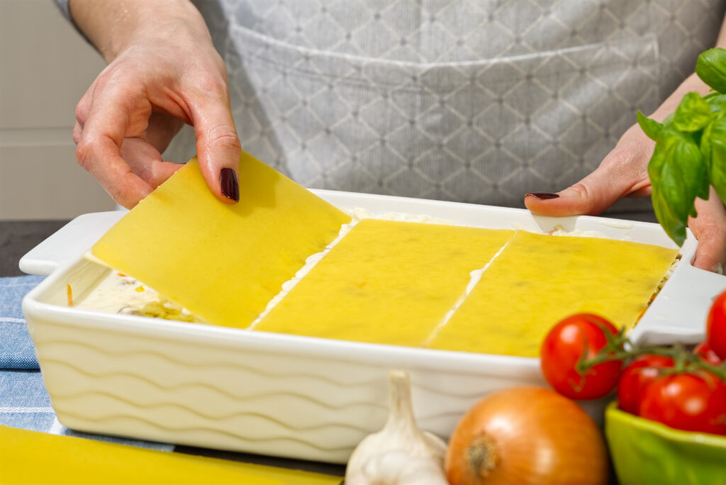 Hands layering lasagna in a dish, starting with sauce, followed by noodles, and then cheese.